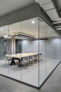Luminous conference zone in the office in a loft style with brick walls and concrete columns. Zone has a large wooden table with gray chairs and glass walls. Above the table there is a projector.